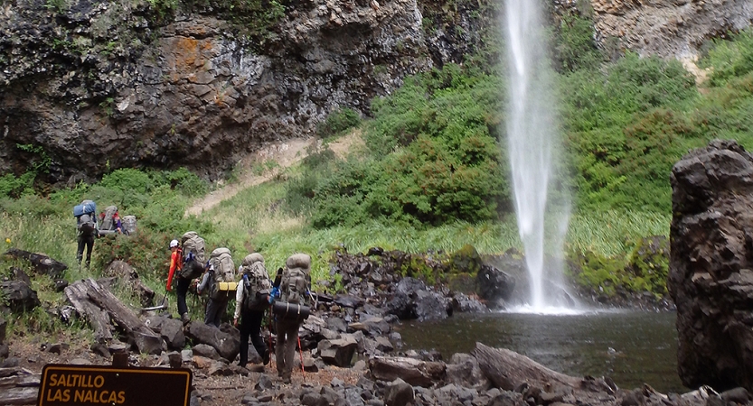 A group of people wearing backpacks hike away from the camera towards a waterfall.